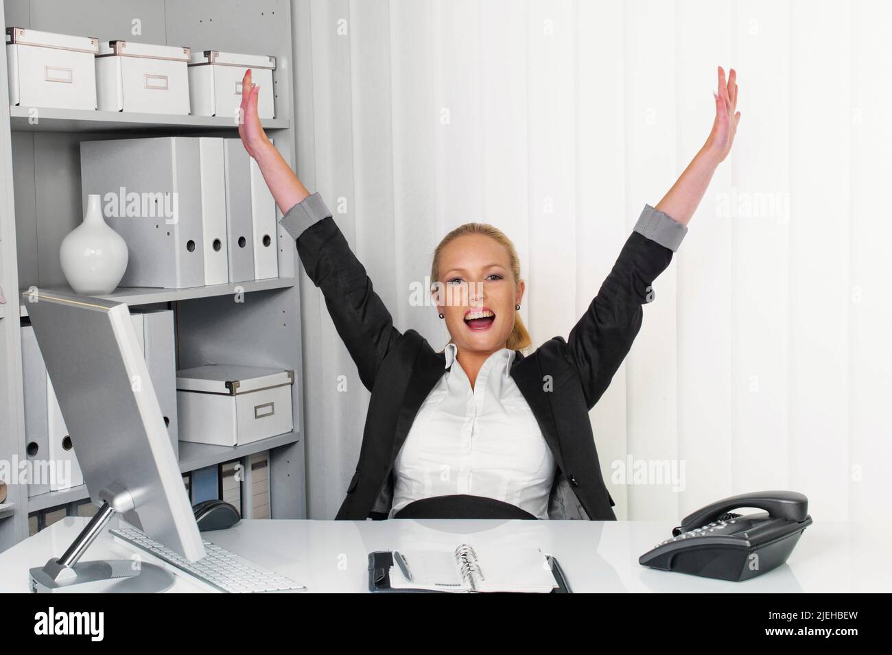 Eine junge erfolgreiche Frau sitzt an ihrem Schreibtisch im Büro und streckt die Armne hoch Stock Photo