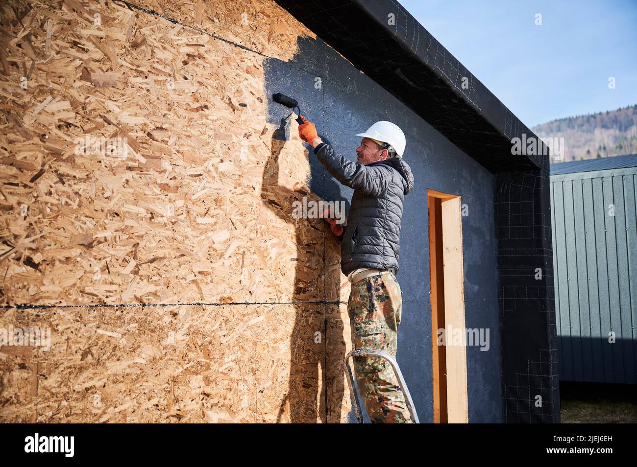 Male painter using paint roller, doing exterior paint work in a black color. Man worker building wooden frame house. Carpentry and construction concept. Stock Photo