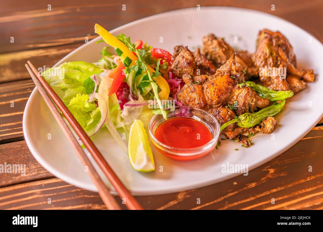 Jamaican jerk chicken plate with vegetables like salada, slices of yellow and red bell peppers, lemon on a plate with chopsticks in a Japanese restaur Stock Photo
