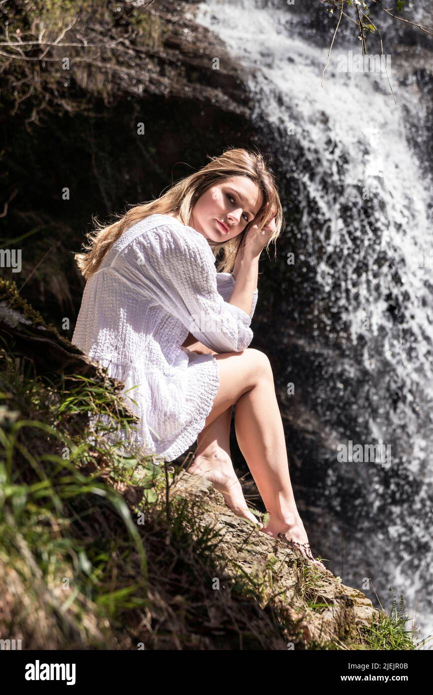 blonde woman in a white dress sitting in the forest in front of a waterfall Stock Photo