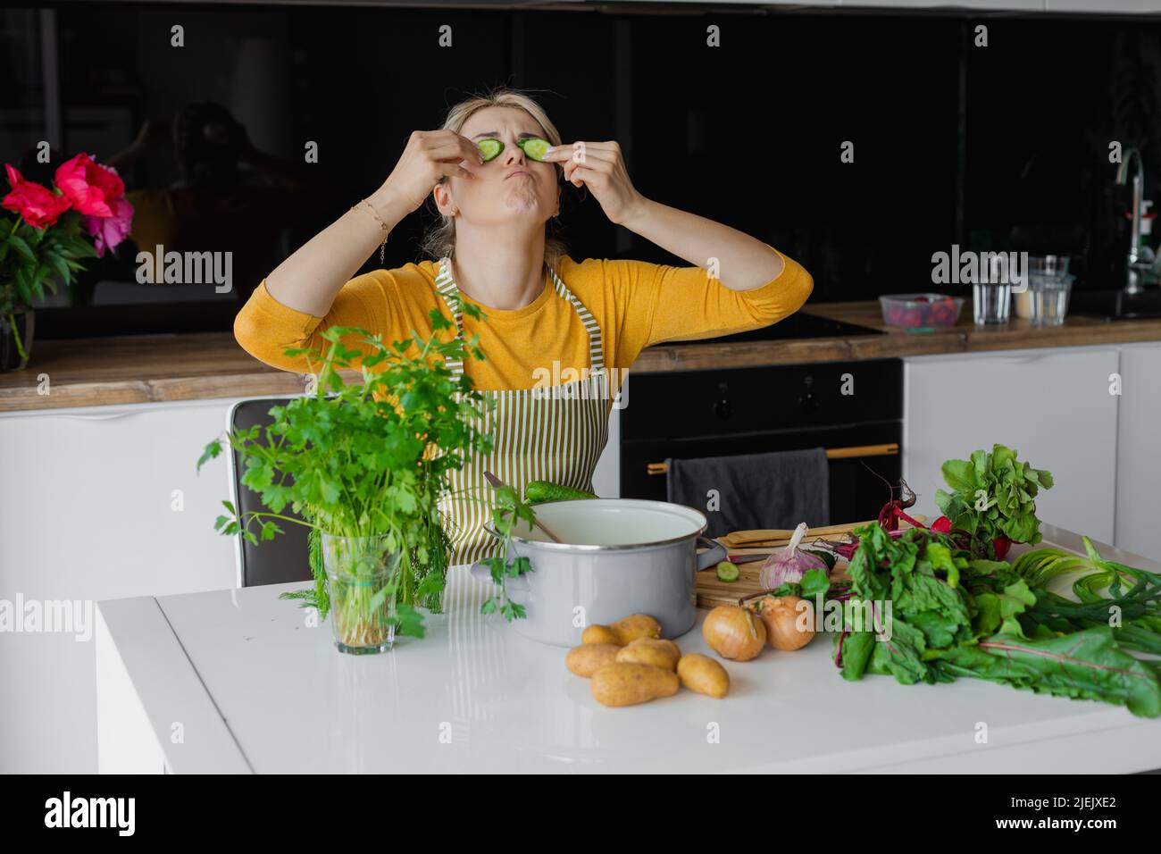 Funny, amusing blonde woman in apron holding cucumber slices on eyes as mask in kitchen. Cosmetology treatment at home Stock Photo