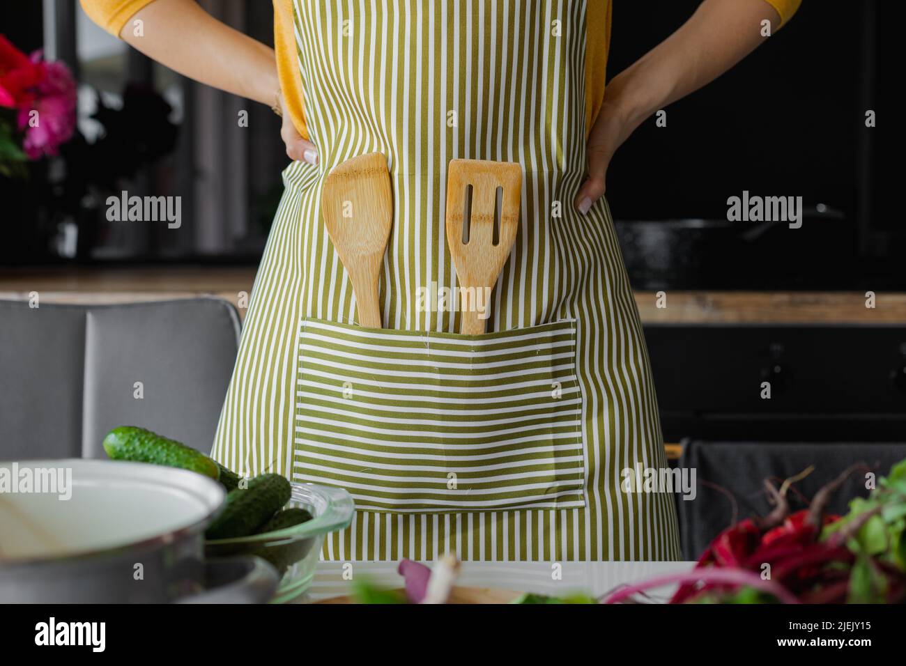 Unrecognizable woman with arms akimbo in striped kitchen apron with wooden spatula utensil. Chef cook fresh food at home Stock Photo