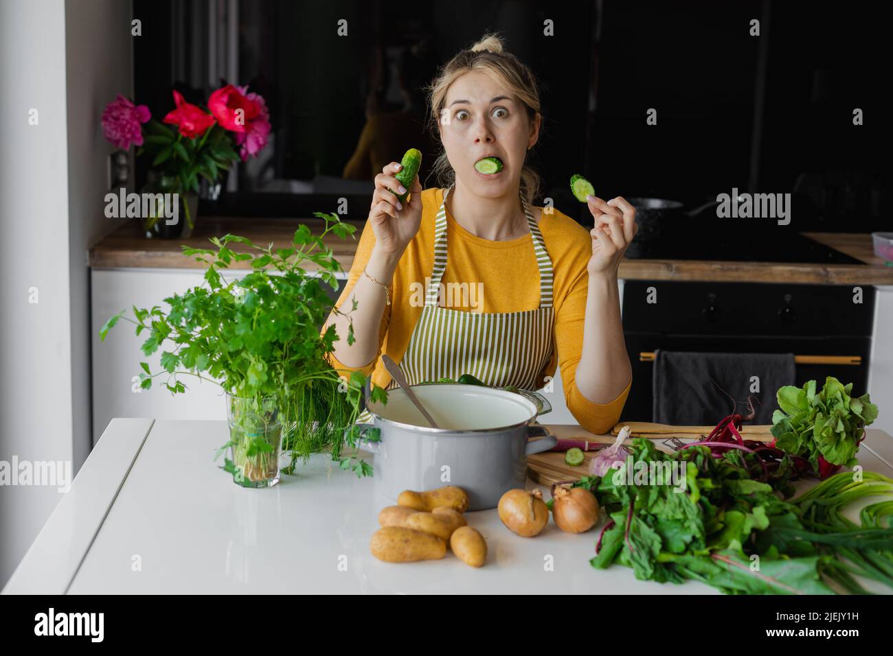 Funny and playful blonde woman looking at camera with cucumber in mouth, chopping fresh vegetables for soup and salad Stock Photo