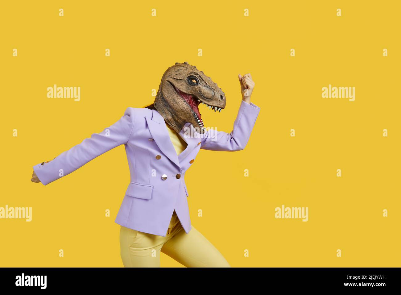 Crazy woman with with t-rex mask threatening someone with her fist, copy space for text on yellow background . Studio shot, portrait. Absurd and funny Stock Photo