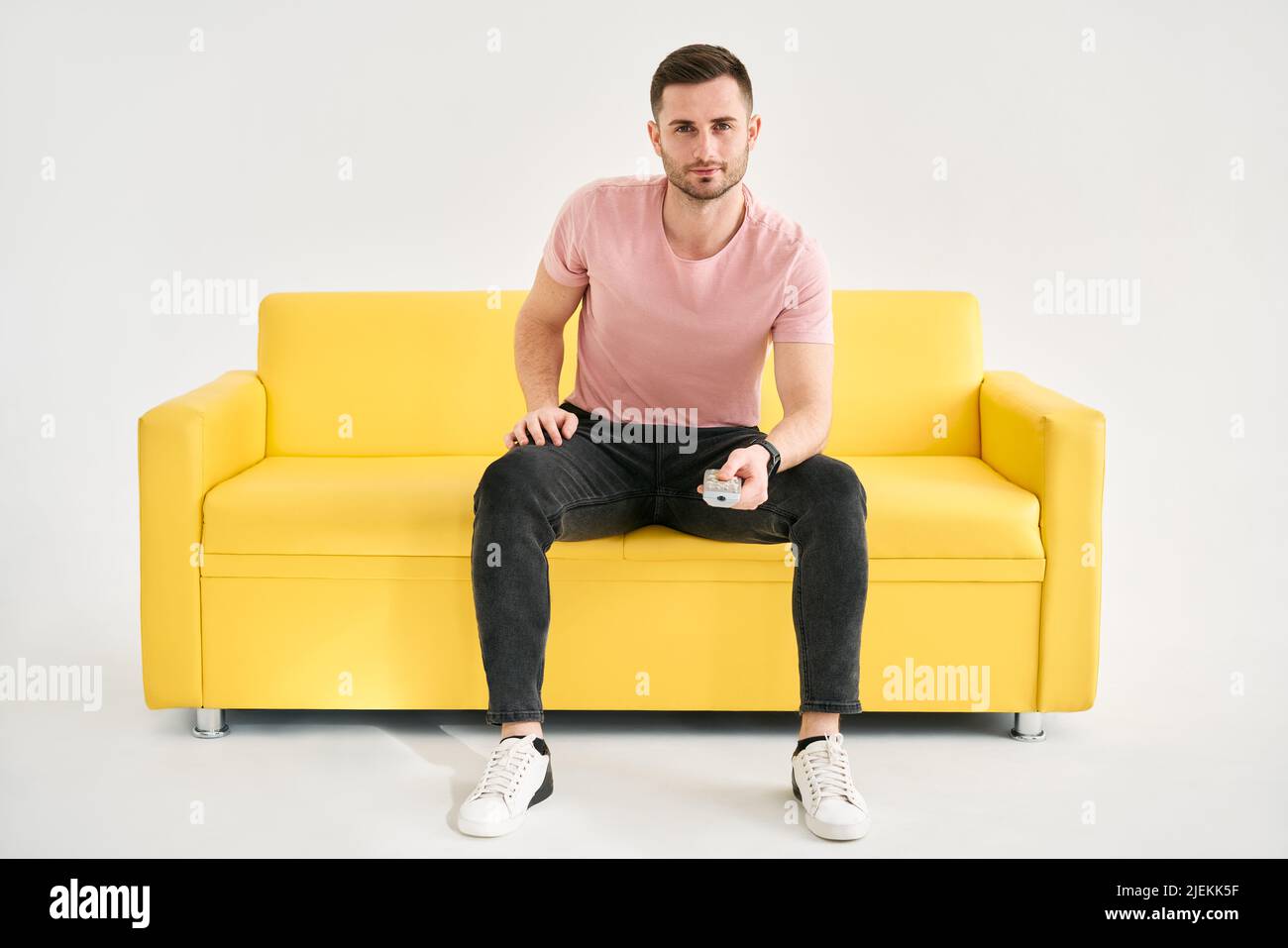 Young man watching TV with remote control sitting on comfort sofa over white background. Male looking to camera holding TV remote control Stock Photo