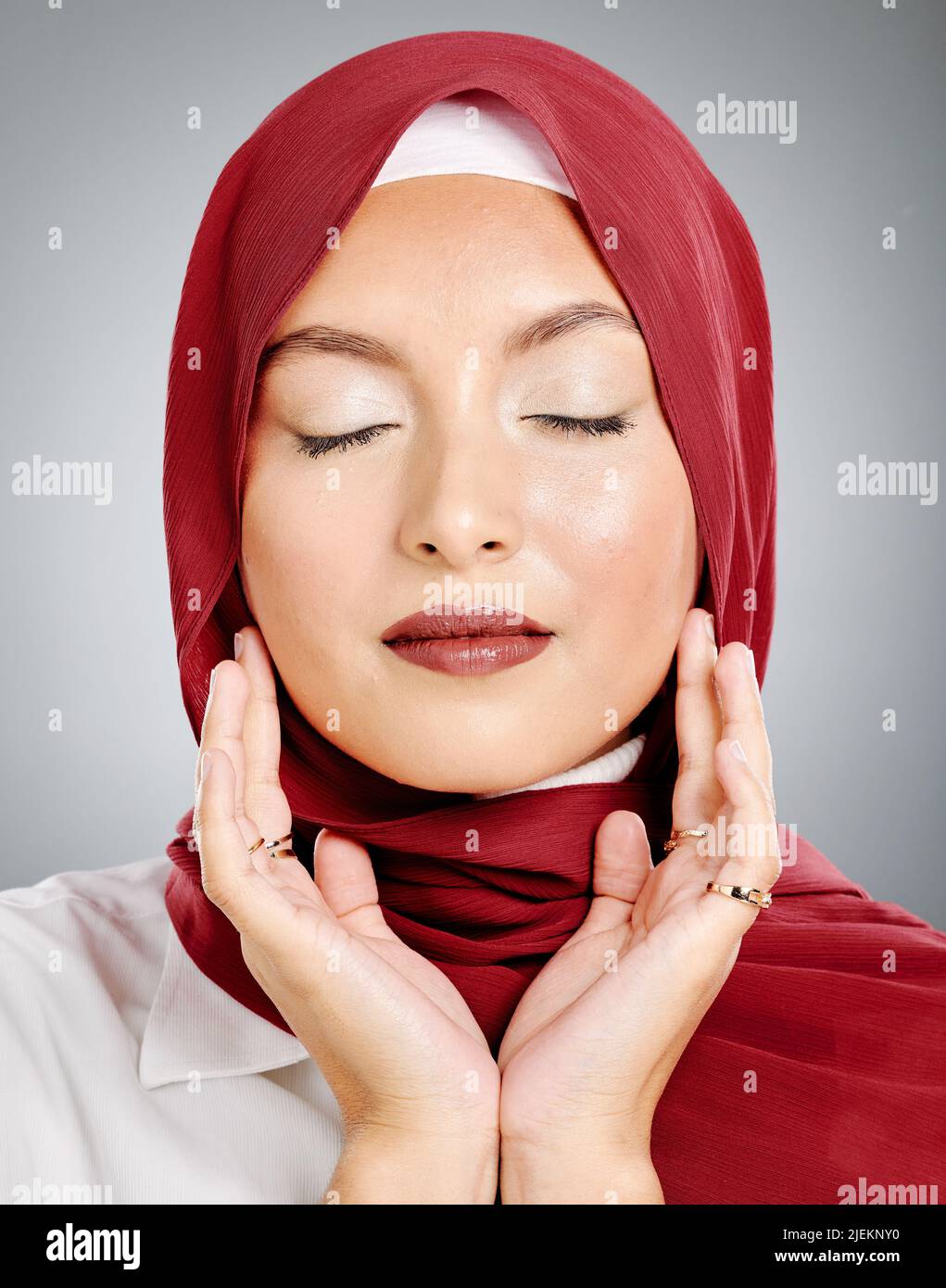 One beautiful young muslim woman with eyes closed wearing red headscarf and lipstick against grey studio background. Modest arab female wearing makeup Stock Photo