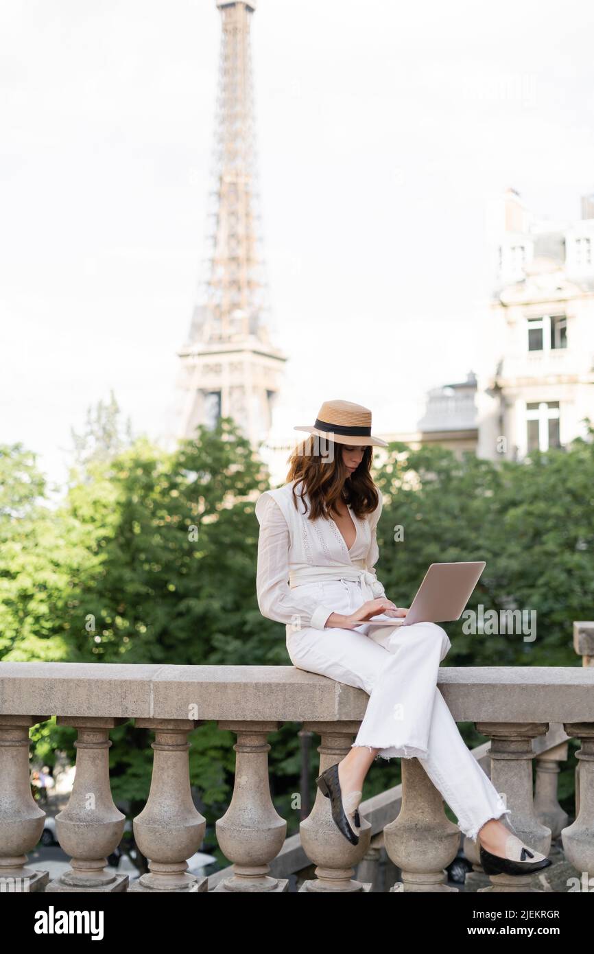 Stylish woman in sun hat using laptop on urban street with Eiffel tower ...