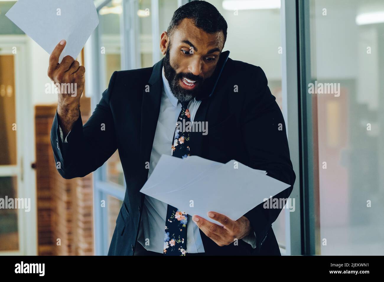 Mid adult bearded black man Entrepreneur Businessman wearing suit holding papers and talking on smartphone stressed walking in office shot Stock Photo
