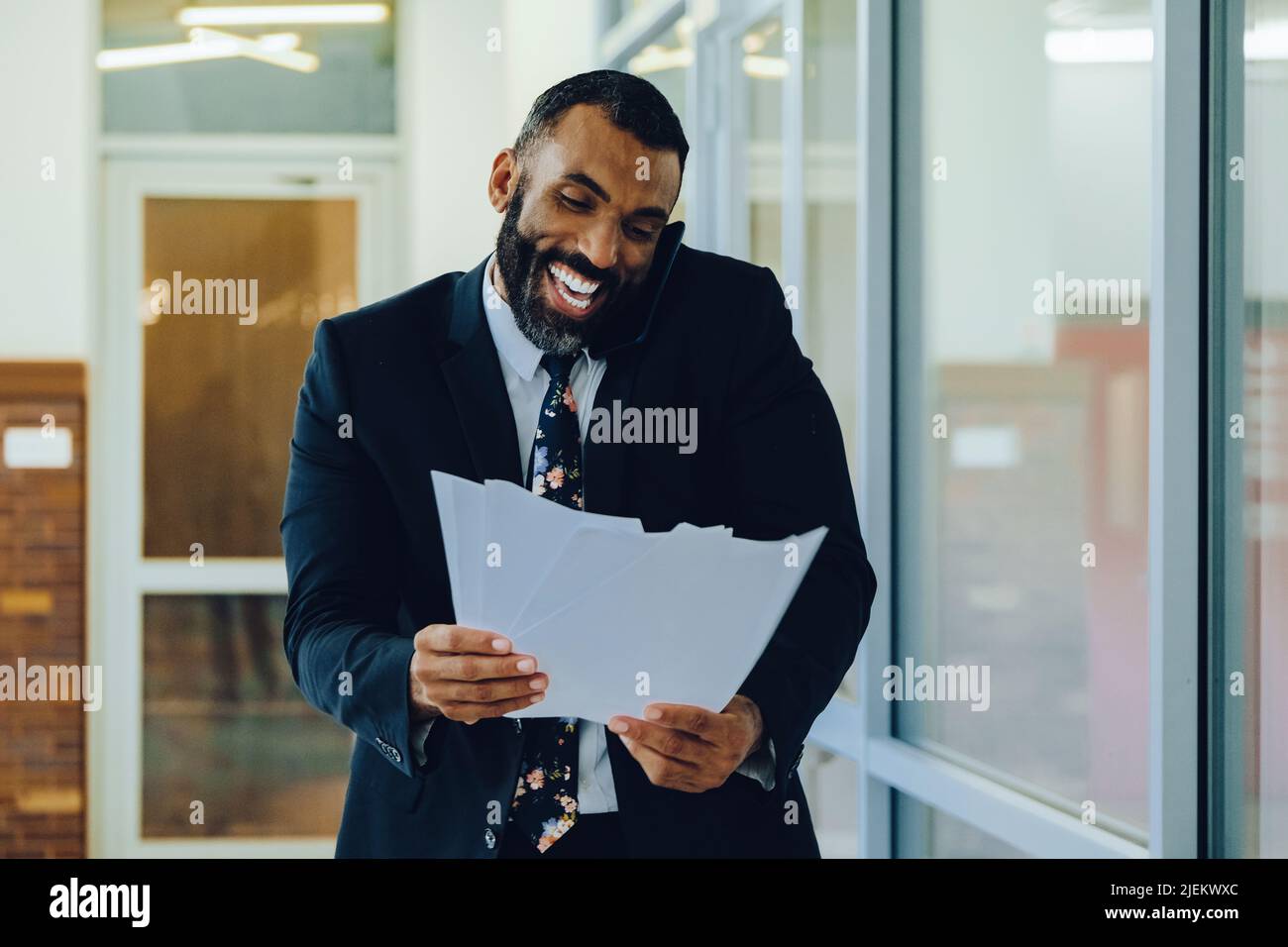 Mid adult bearded black man Entrepreneur Businessman wearing suit holding papers and talking on smartphone laughing walking in office shot Stock Photo