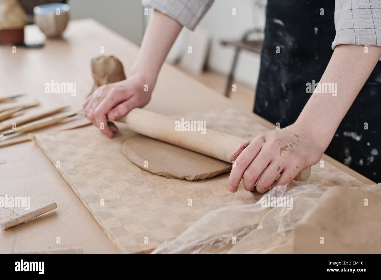 Hands of young craftswoman with wooden rolling pin flattening clay on board while working over new clay products for sale in workshop Stock Photo