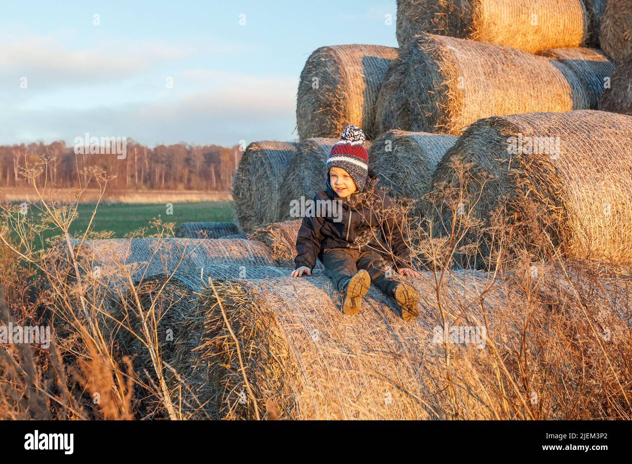 In the background of golden hay bales stack, and rural countryside landscape, poses a cute smiling boy in autumn clothes and a knitted hat. Stock Photo