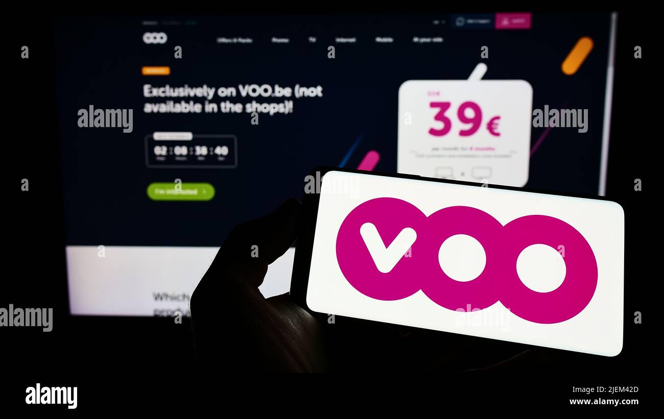 Person holding mobile phone with logo of Belgian telecommunications company Voo SA on screen in front of business web page. Focus on phone display. Stock Photo