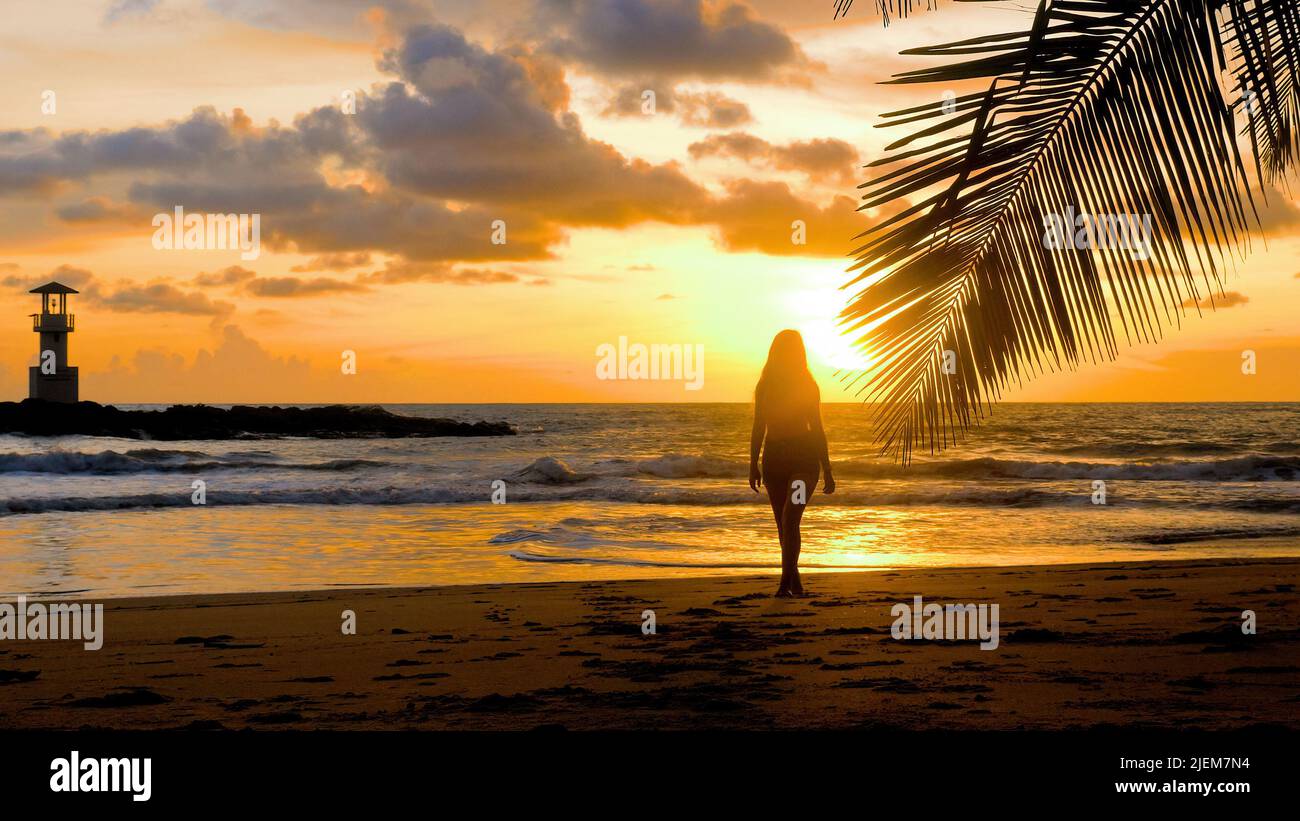Young happy woman walking barefoot on sandy beach to palm tree branch with sea, sunset sun and lighthouse on background. Girl silhouette enjoy summer Stock Photo