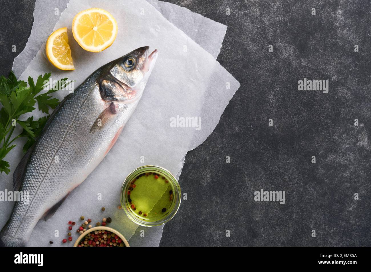 Sea Bass raw. Fresh sea fish bass with salt, pepper, parsley, olive oil and lemon on parchment paper on a dark concrete rustic background. Food cookin Stock Photo