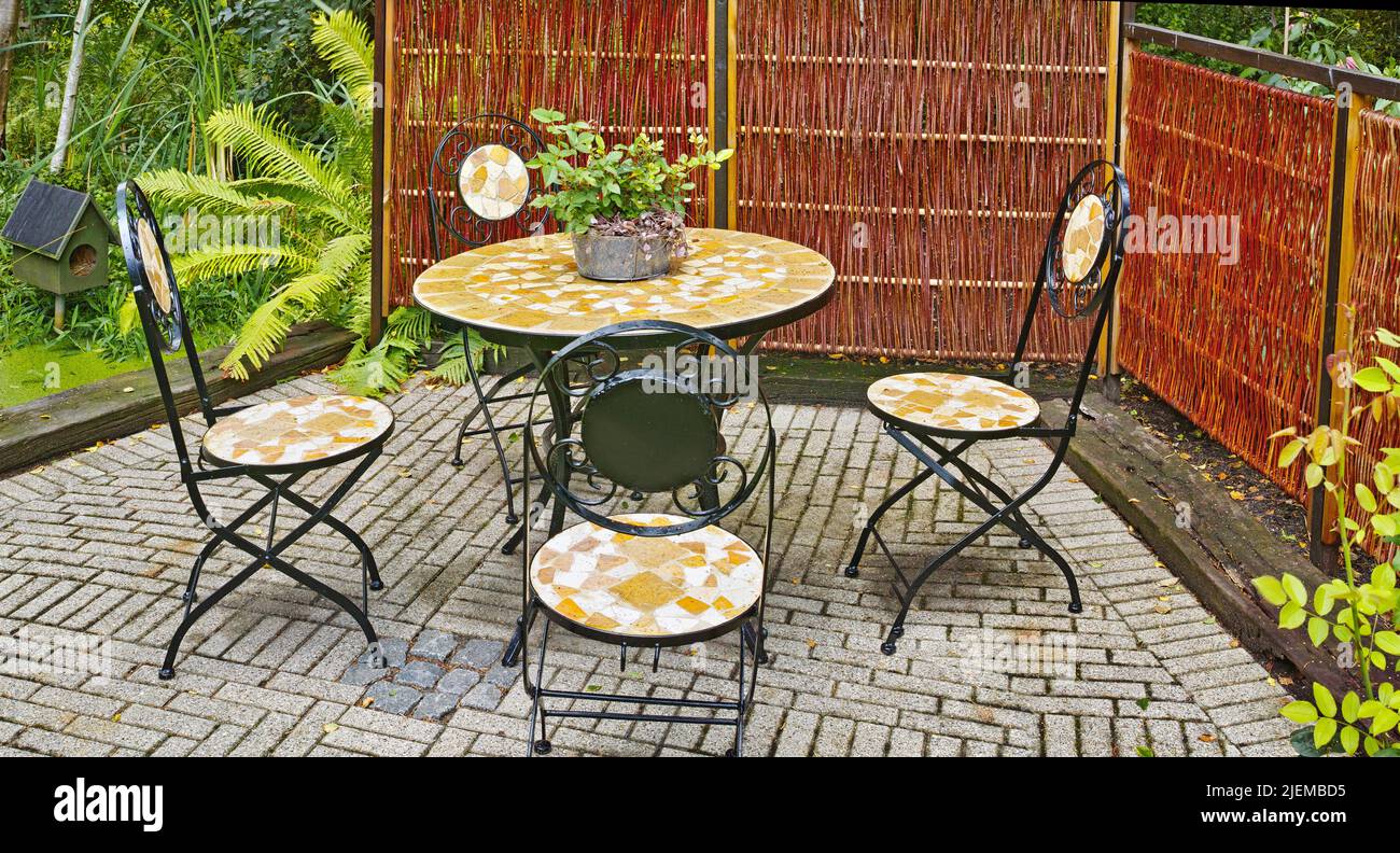 Set of outdoor dining furniture. Mosaic tile and iron chairs and table on a grey brick patio in a lush garden. Cozy enclosed relaxation area outside Stock Photo