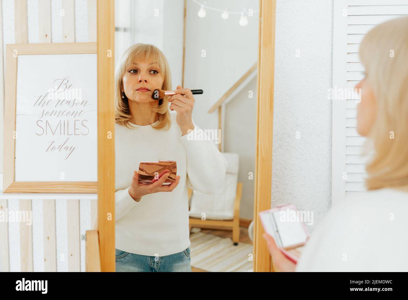 Beautiful smiling mature senior caucasian woman with blonde hair standing near mirror with make up palette and brush, doing makeup and looking at Stock Photo