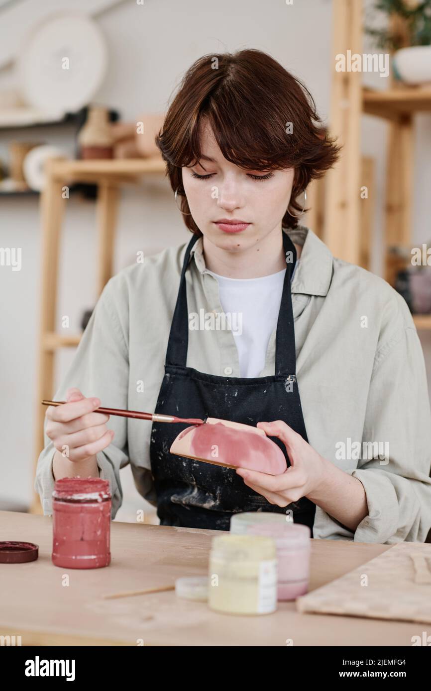 Young serious skilled female artisan with clay bowl and paintbrush painting handmade earthenware while sitting by workplace Stock Photo