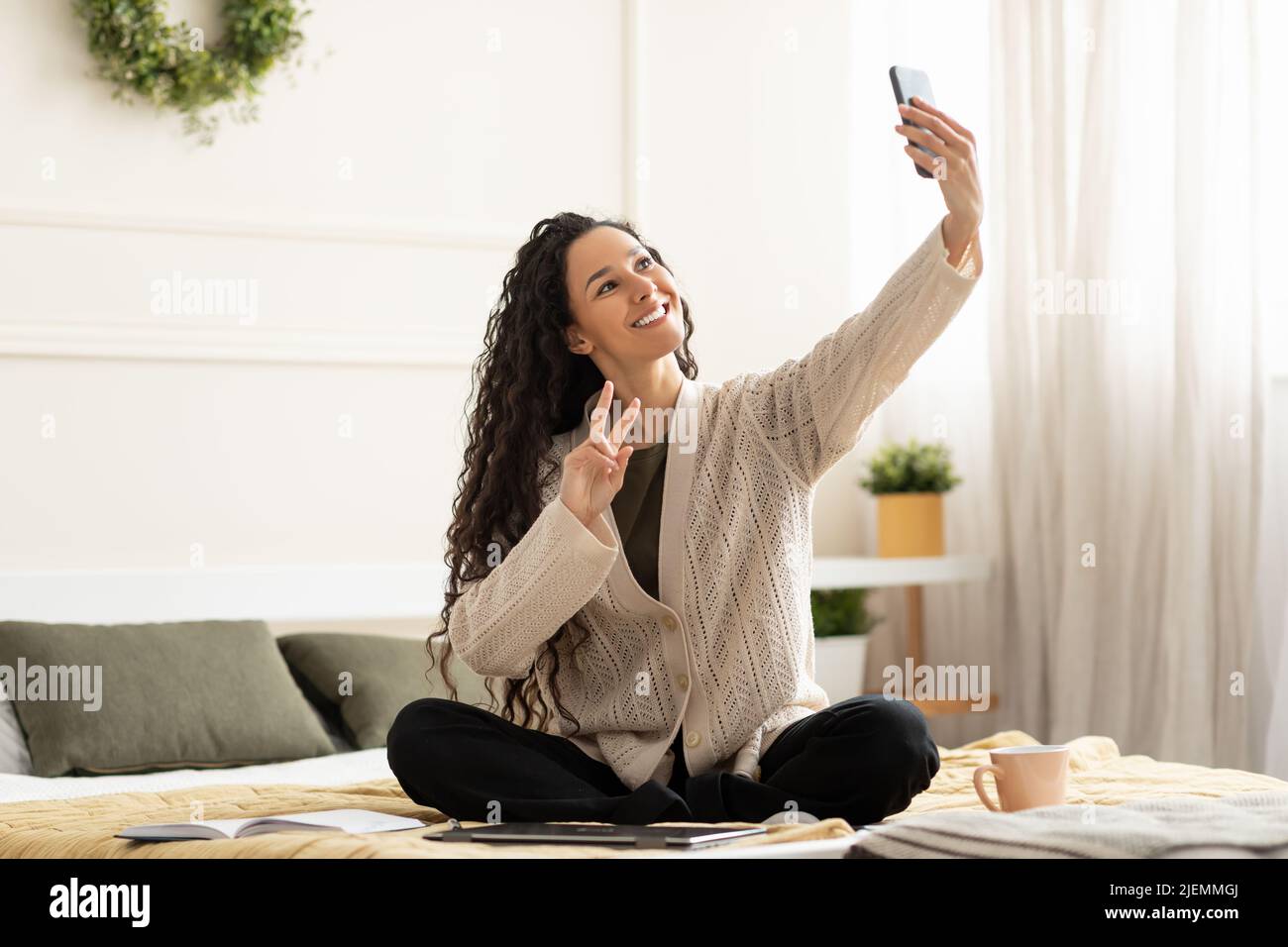 Happy millennial female vlogger taking selfie, live streaming on mobile phone, sitting on bed at home Stock Photo