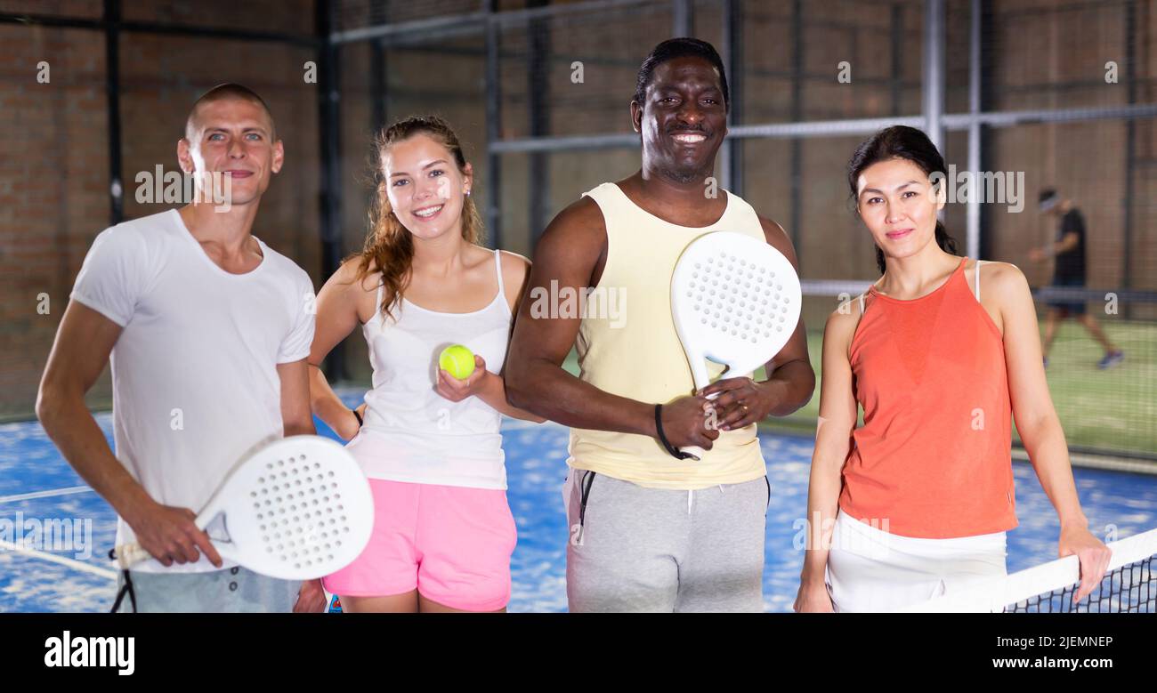 Smiling men and women with rackets and balls posing on indoor padel court Stock Photo