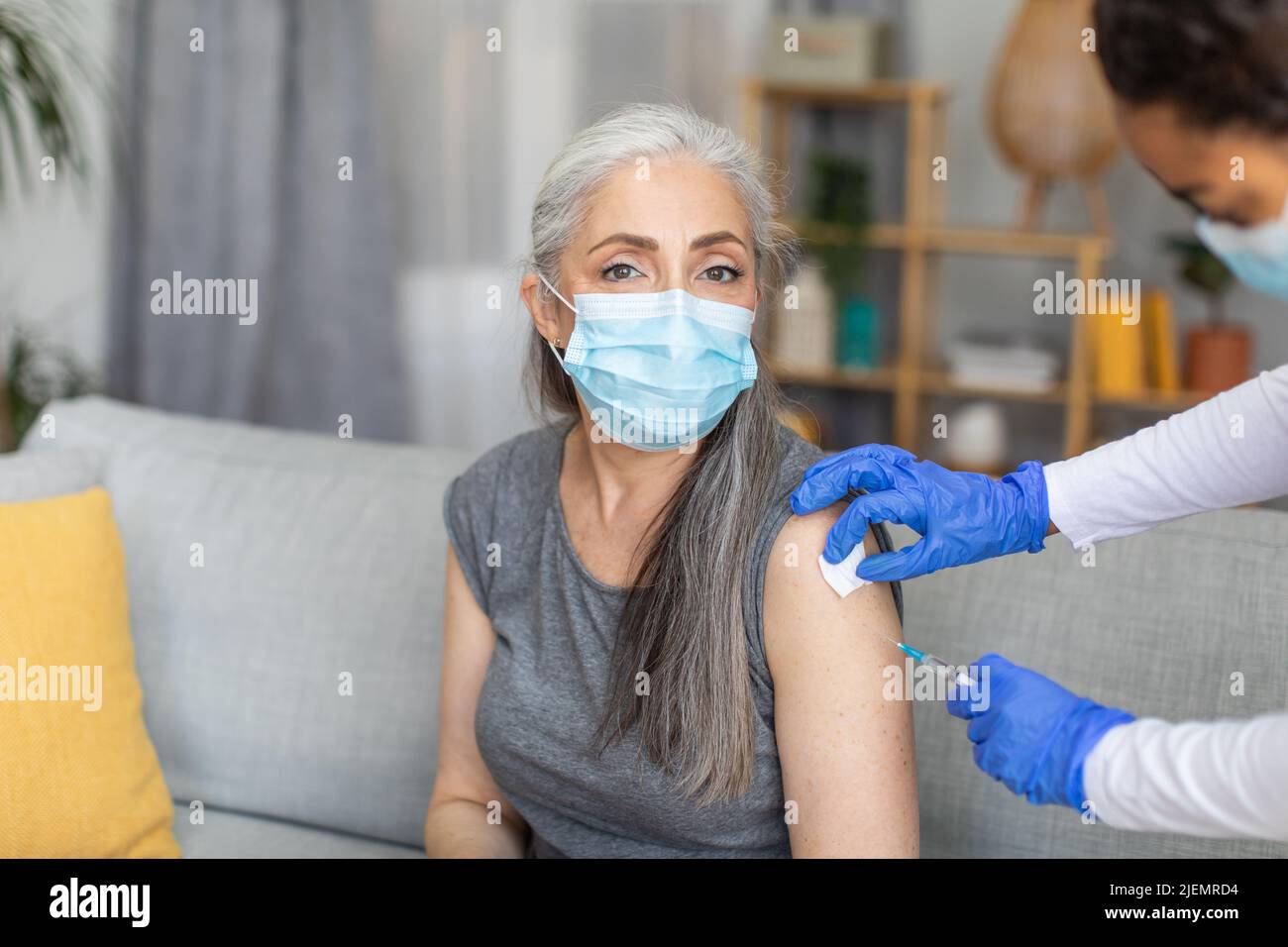 Doctor in protective gloves makes injection of european old woman in mask with gray hair in living room Stock Photo
