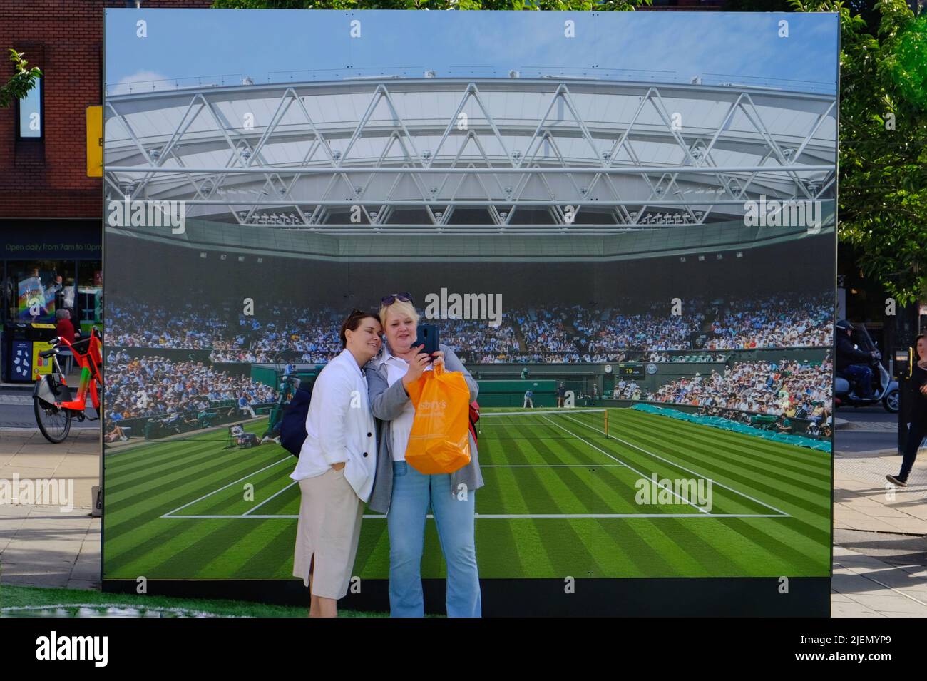 London, UK, 27th June, 2022. Two women take selfies in front of a Centre Court backdrop. Tennis themed shop displays and in Wimbledon Village and the main town centre can be seen as the 2022 tennis tournament commences. This year the ground returns to full capacity after the Covid pandemic. Credit: Eleventh Hour Photography/Alamy Live News Stock Photo