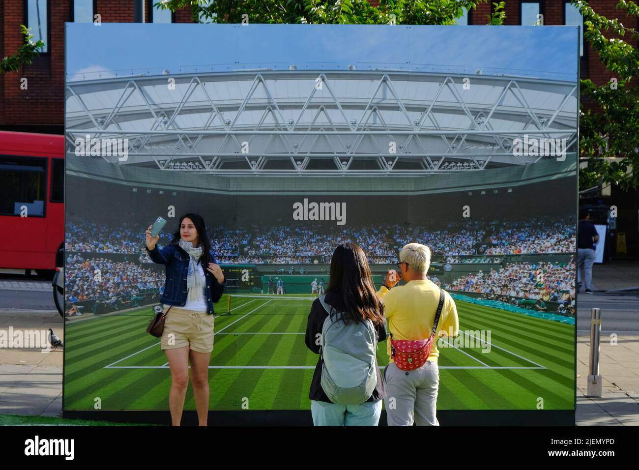 London, UK, 27th June, 2022. A woman takes a sefie in front of a Centre Court photo backdrop. Tennis themed shop displays and in Wimbledon Village and the main town centre can be seen as visitors are welcomes for  the 2022 tennis tournament. This year the ground returns to full capacity after the Covid pandemic. Credit: Eleventh Hour Photography/Alamy Live News Stock Photo