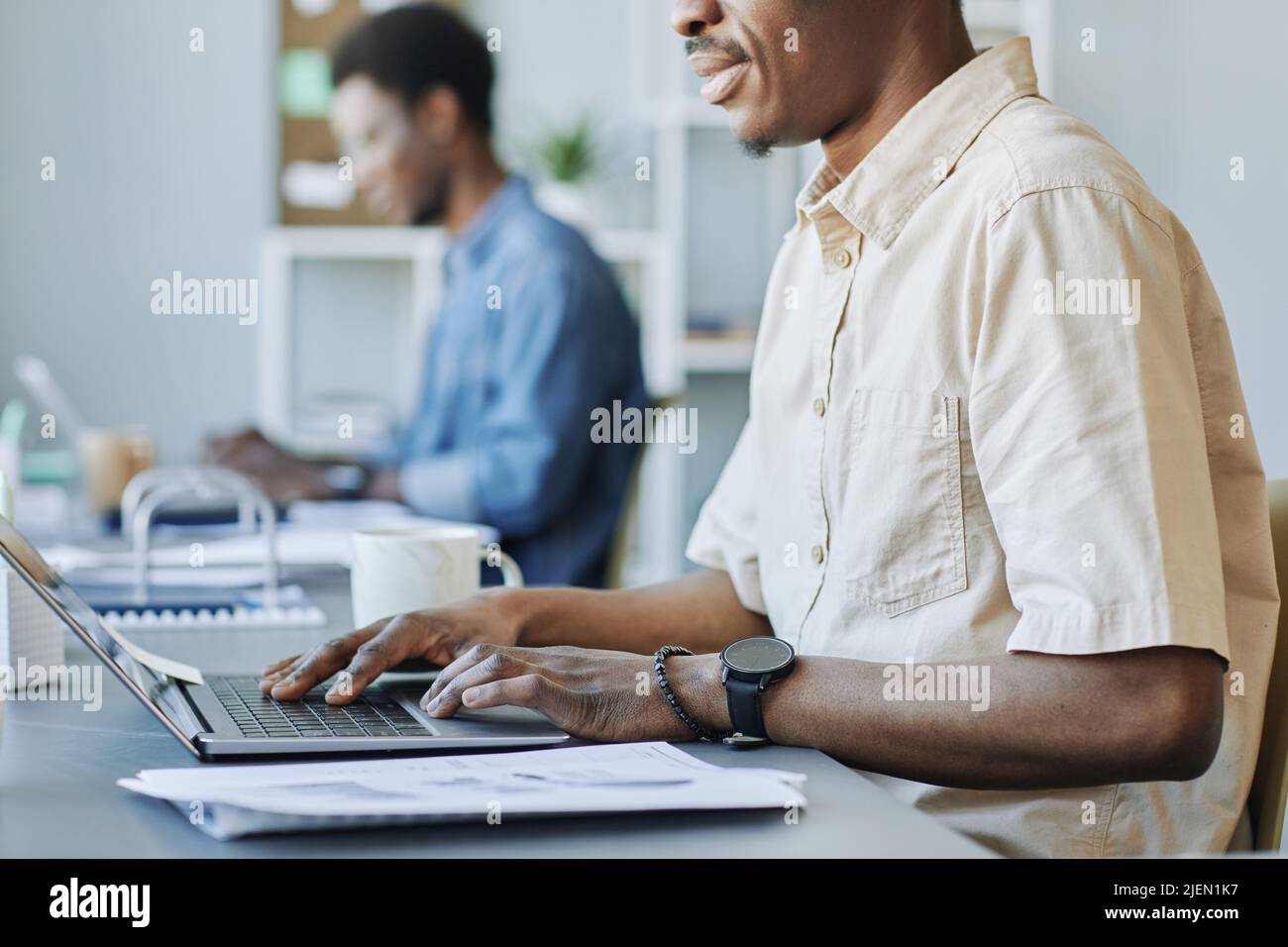 Cropped side view shot of adult black man typing at laptop while working in office and holding cup of coffee, copy space Stock Photo