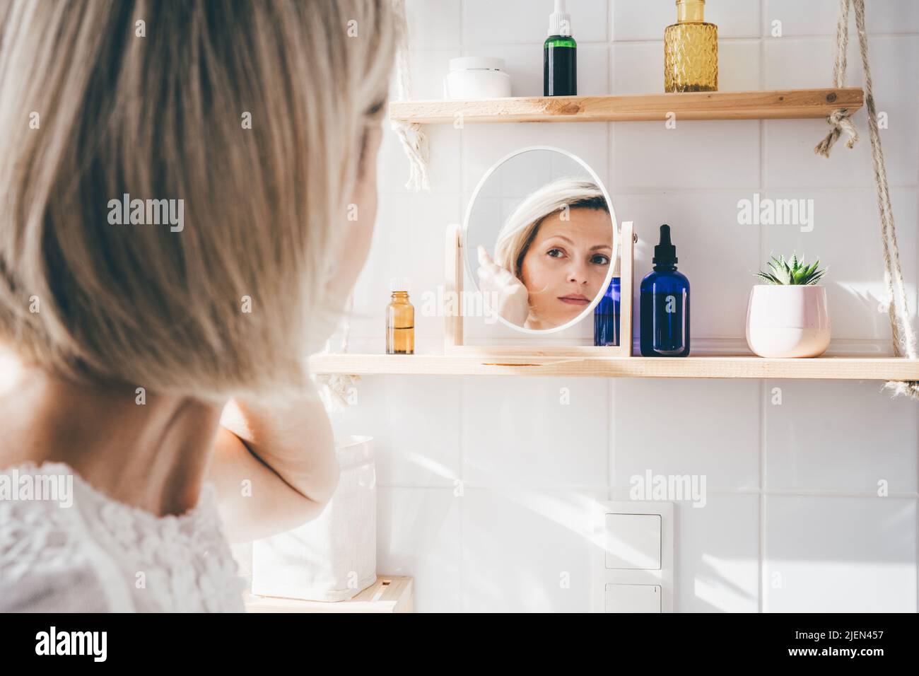 Beautiful middle-aged woman looking in the mirror in white eco friendly bathroom. Wooden shleves and reusable cosmetics bottles. Wellnes concept Stock Photo