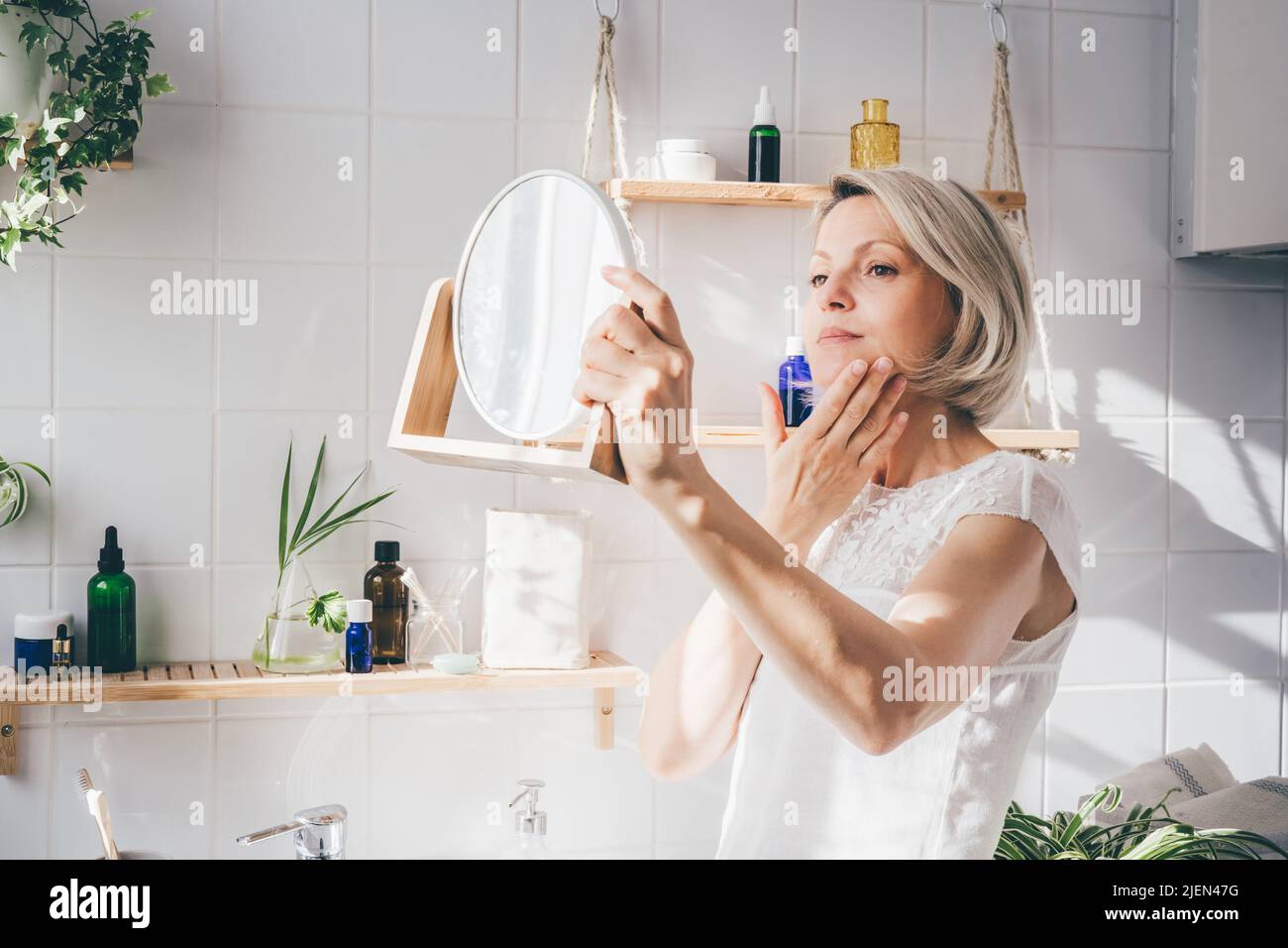 Beautiful middle-aged woman taking care of her face and looking in the mirror. White eco-friendly bathroom interior. Natural light. Wellness Stock Photo
