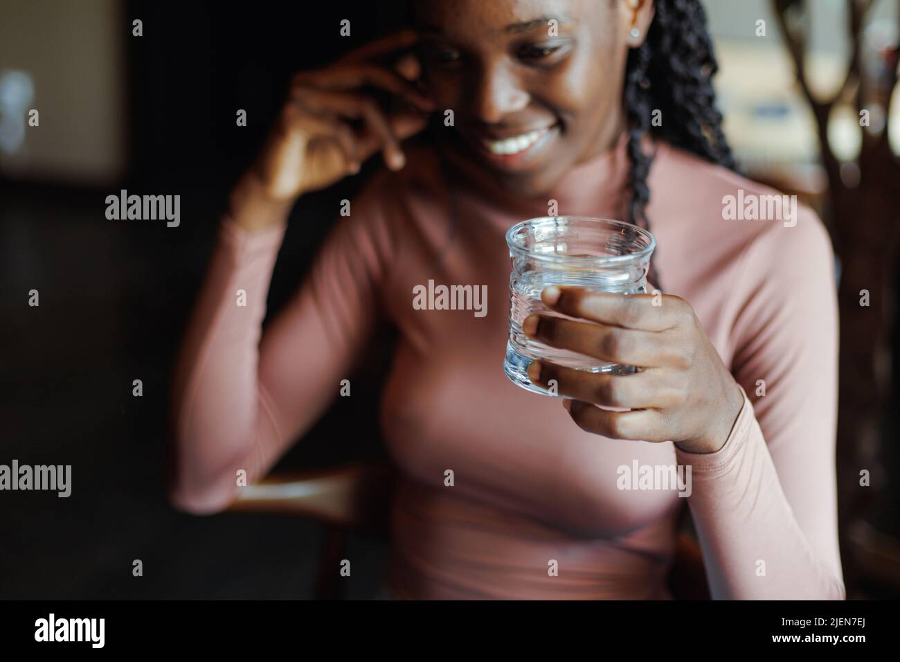 Close-up of young smiling cute African-American woman sitting at beige table, holding glass of water, looking aside. Stock Photo