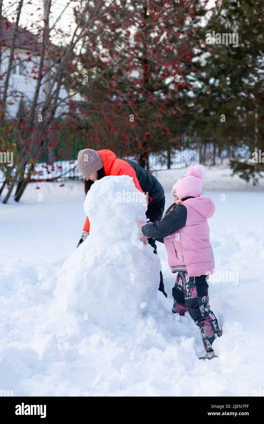 Woman with little daughter on snow making snowman with hands together in evening with rowan and fir trees and fence in background. Winter family time Stock Photo