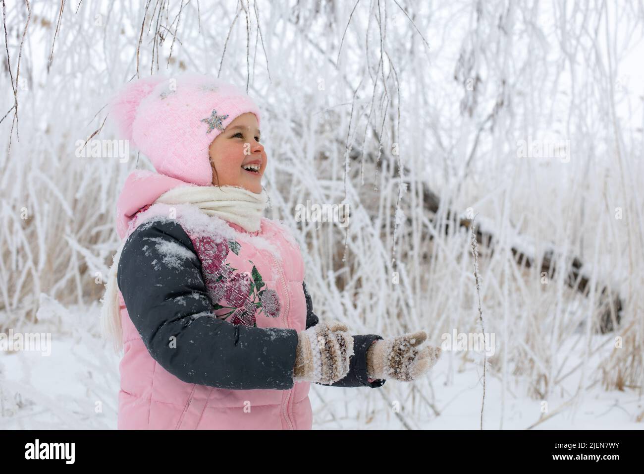 Little girl playing with snow outside dressed in warm winter clothes looking up and smiling with snowy tree branches in background. Winter walk in Stock Photo