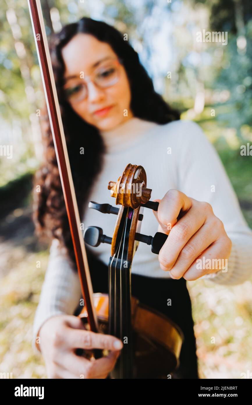 Curly brunette woman with glasses tuning her violin outside in the woods. Vertical. Selective focus. Stock Photo
