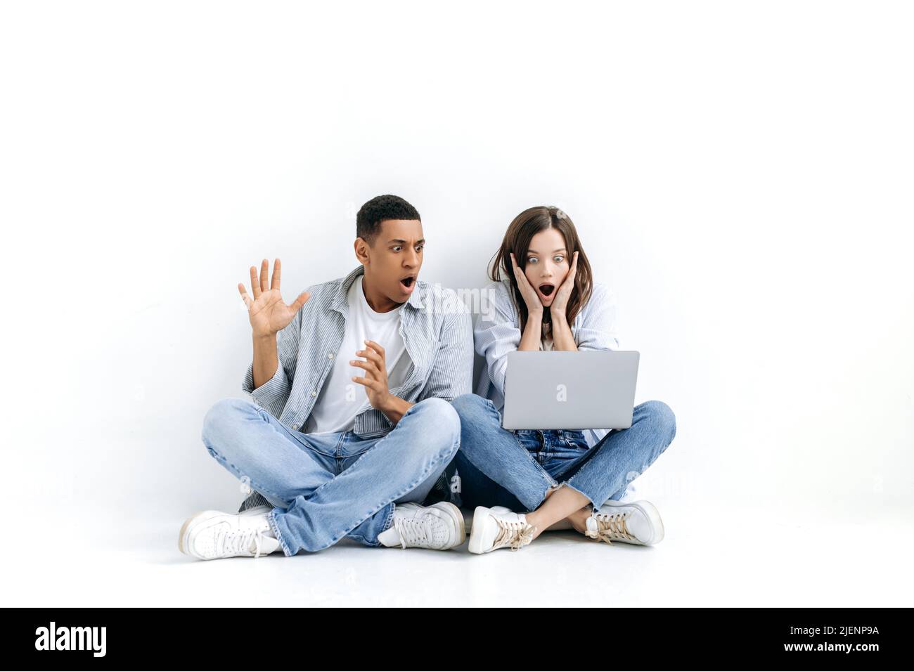 Amazed stunned young multiracial couple, in casual wear, sitting on floor over isolated white background with laptop, looking at a screen in surprise, saw unexpected news, big discount in online store Stock Photo