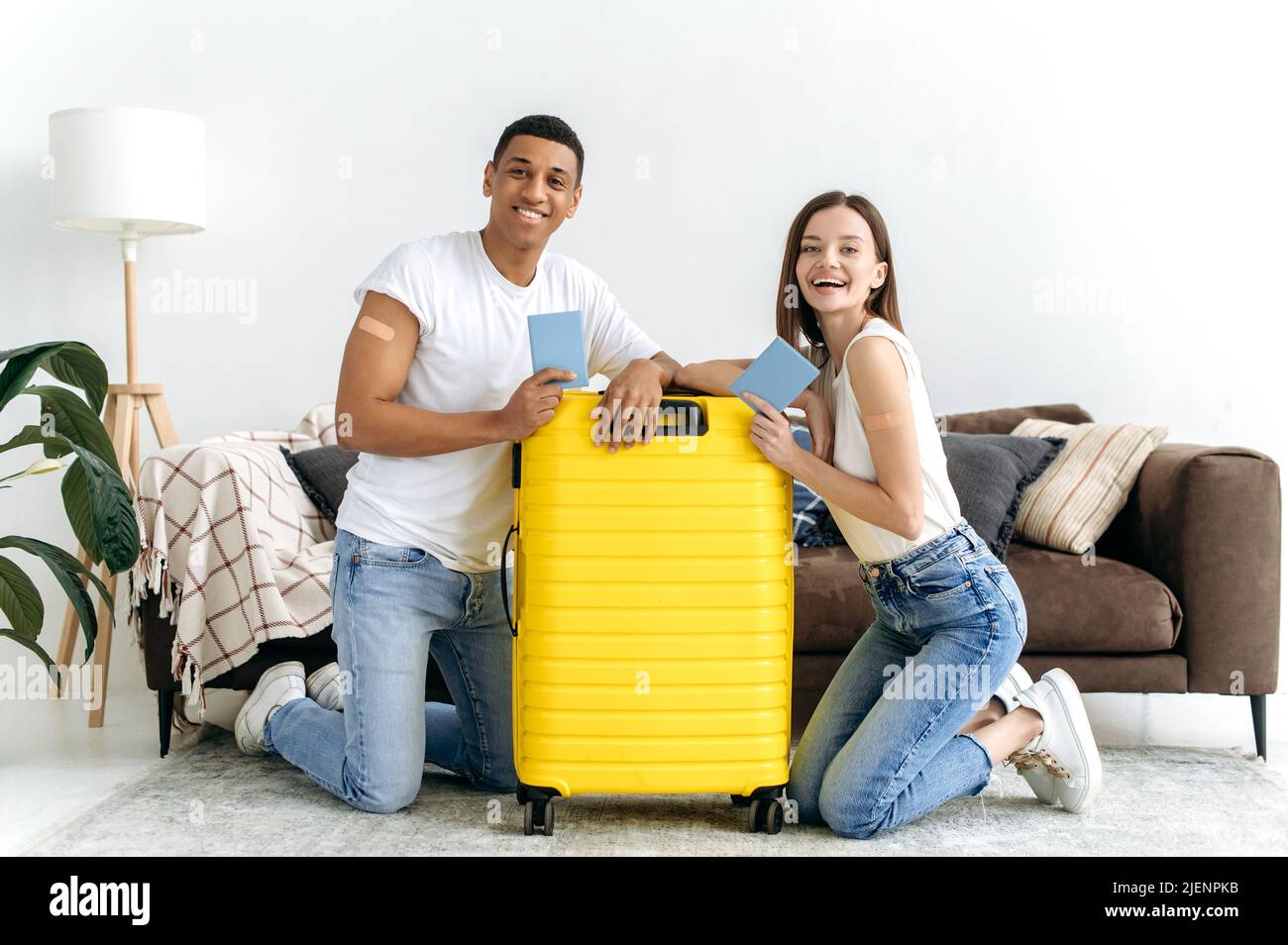 Long-awaited vacation, travel. Happy multiracial couple, got the vaccine and going on a trip, sit on the floor in the living room near the yellow suitcase, hold their passports, look at camera, smile Stock Photo