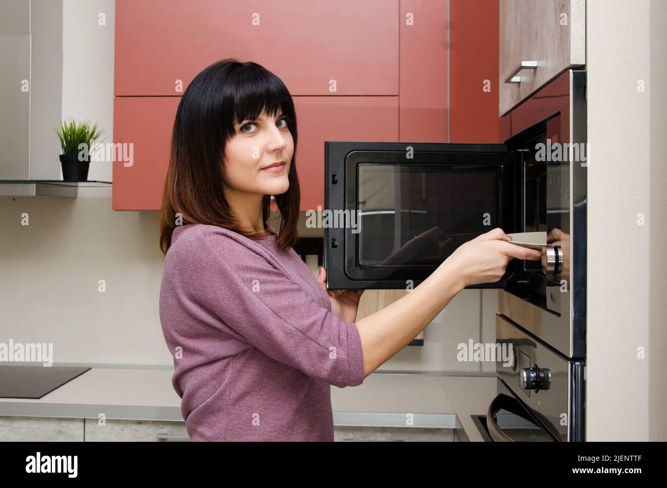 A young woman looks at the camera and takes out a plate from the microwave Stock Photo