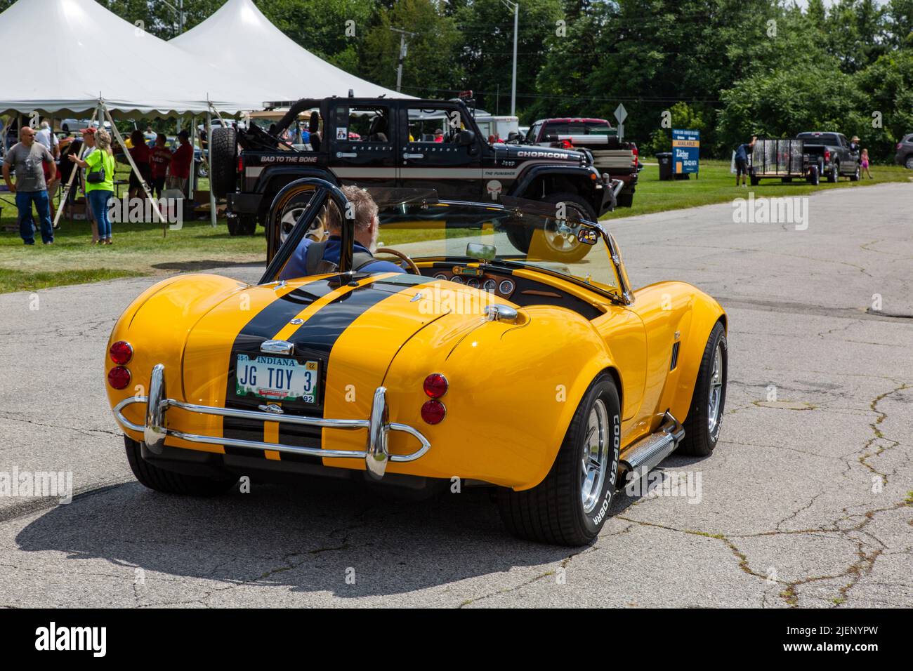 A man drives his yellow AC Cobra replica sports car at a car show in
