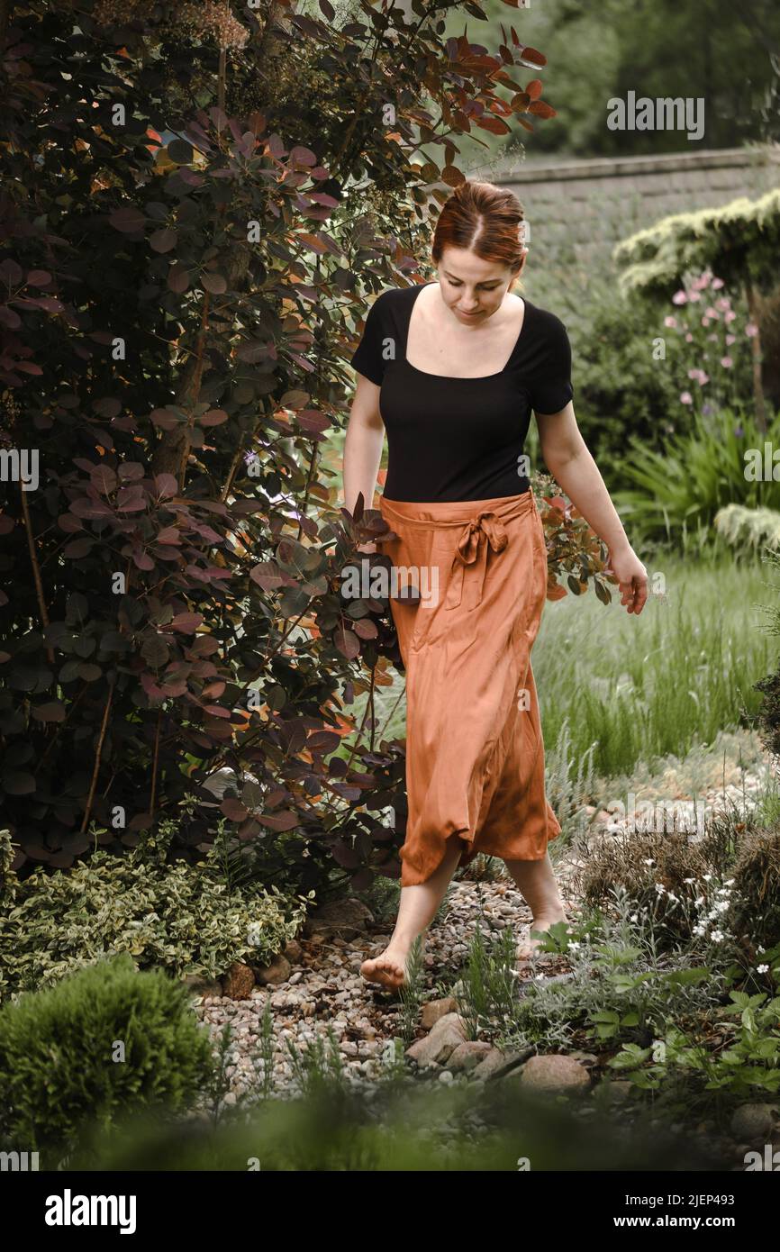 Happy ginger hair woman walking barefoot in the park or garden. Freedom and healthy way of life. Female in her 40s having rest outdoors. People in Stock Photo