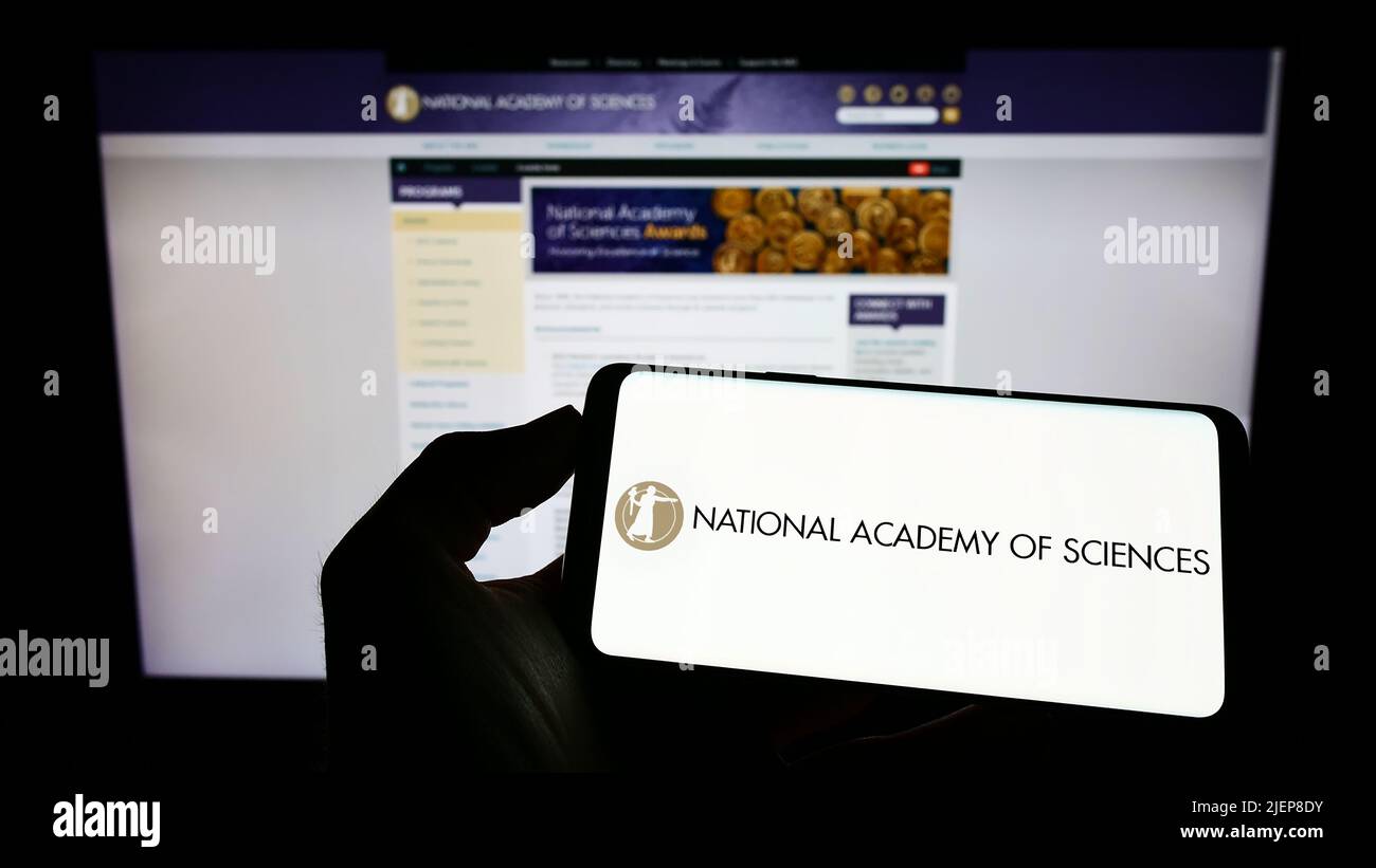 Person holding cellphone with logo of National Academy of Sciences (NAS) on screen in front of webpage. Focus on phone display. Stock Photo