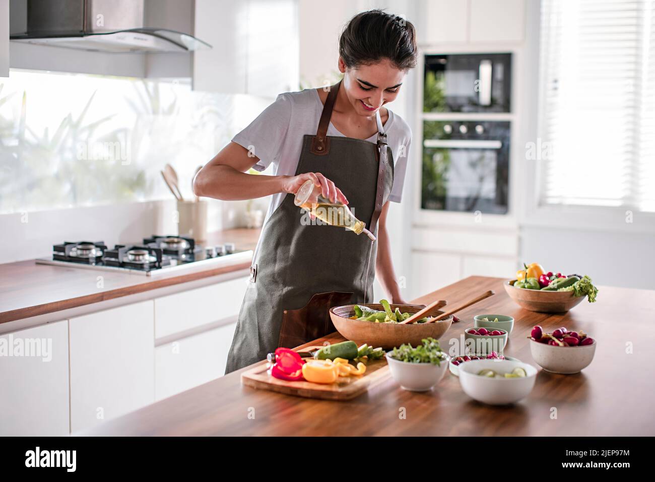 Young woman pouring olive oil on the vegetable salad in the kitchen Stock Photo