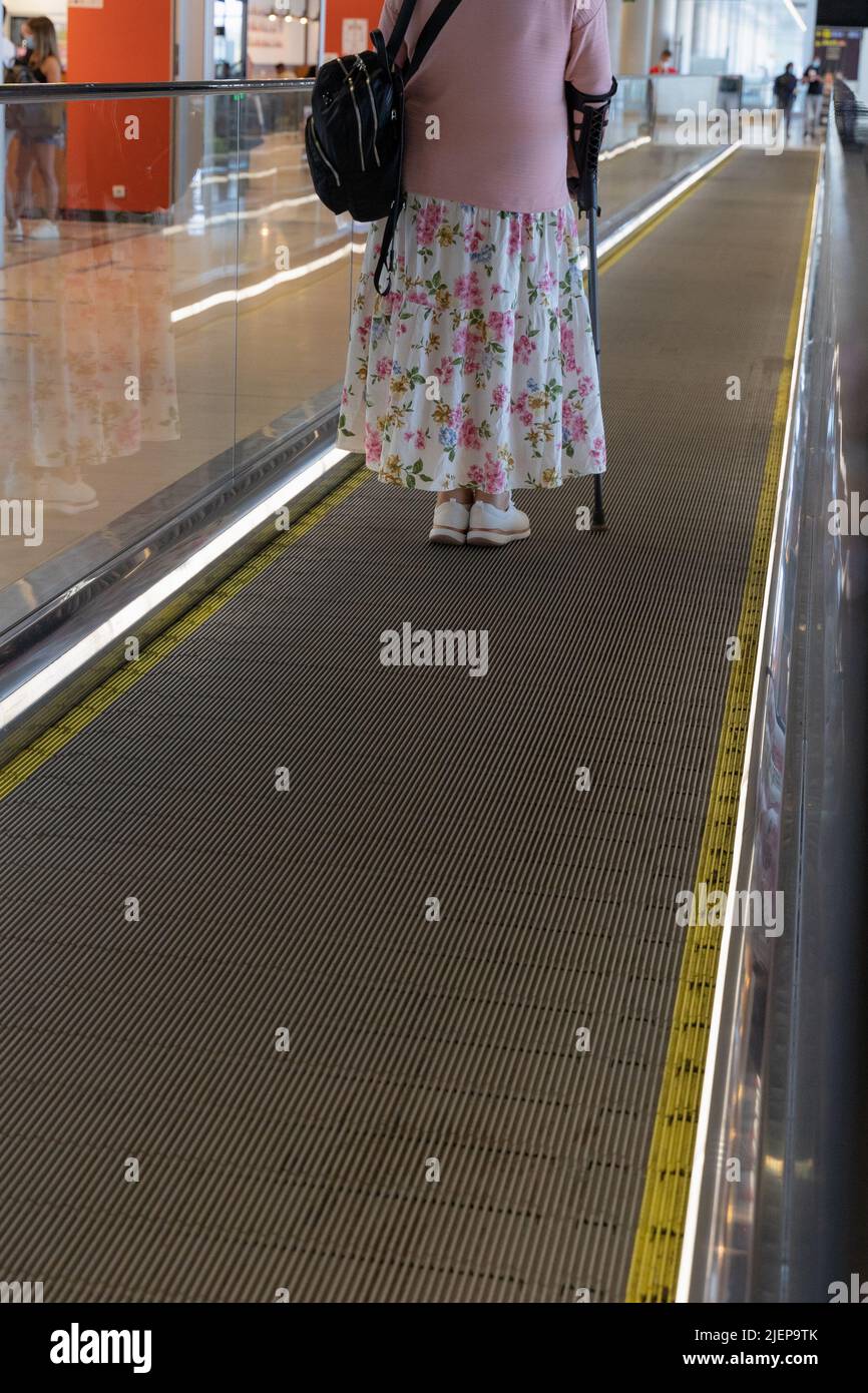 Anonymous elder woman with crutch standing on moving walkway at airport. Travel special assistance concept Stock Photo