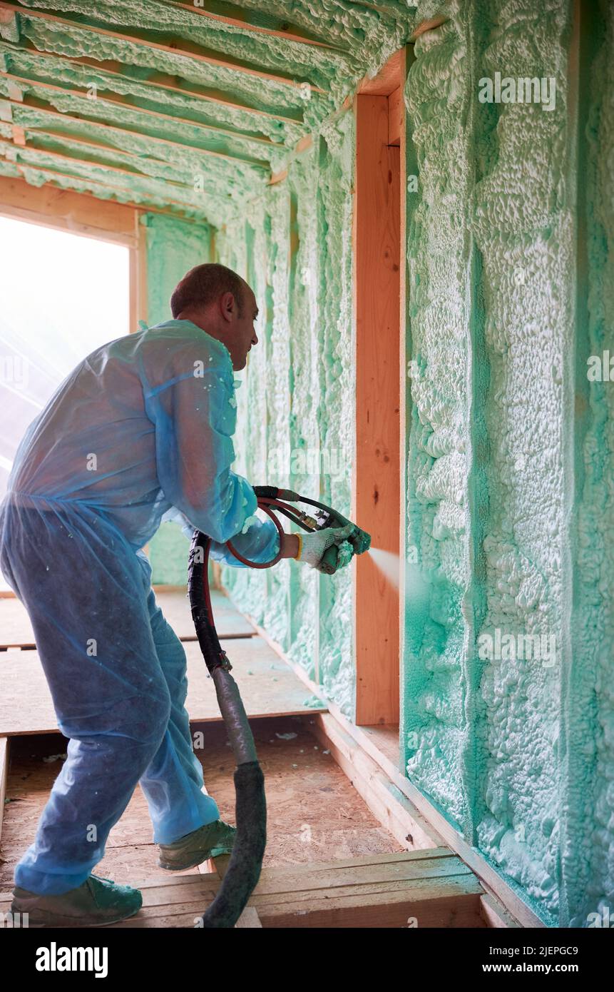 Male builder insulating wooden frame house. Man worker spraying polyurethane foam inside of future cottage, using plural component gun. Construction and insulation concept. Stock Photo
