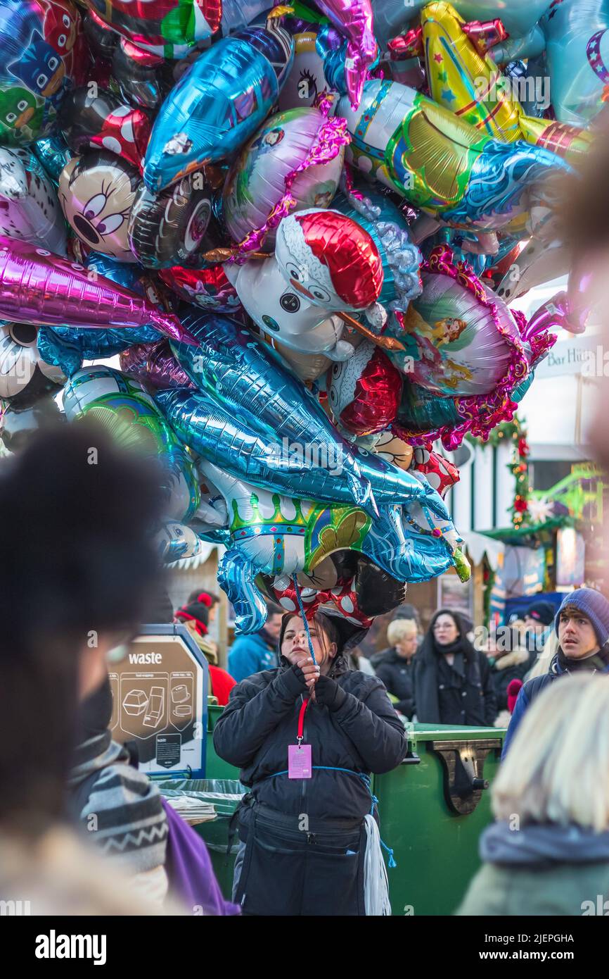 London, UK - 2 December, 2021 - Female street vendor sells multiple cartoon character helium balloons in Christmas funfair winter wonderland Stock Photo