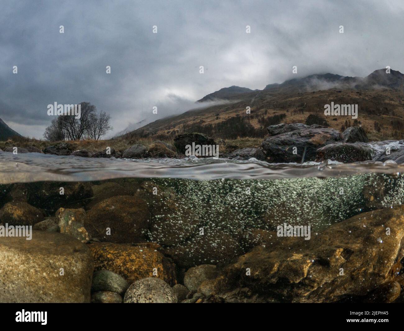 Water flows of the rocks underneath the River Kinglas. Hills, trees and dark stormy clouds are in the background. Stock Photo