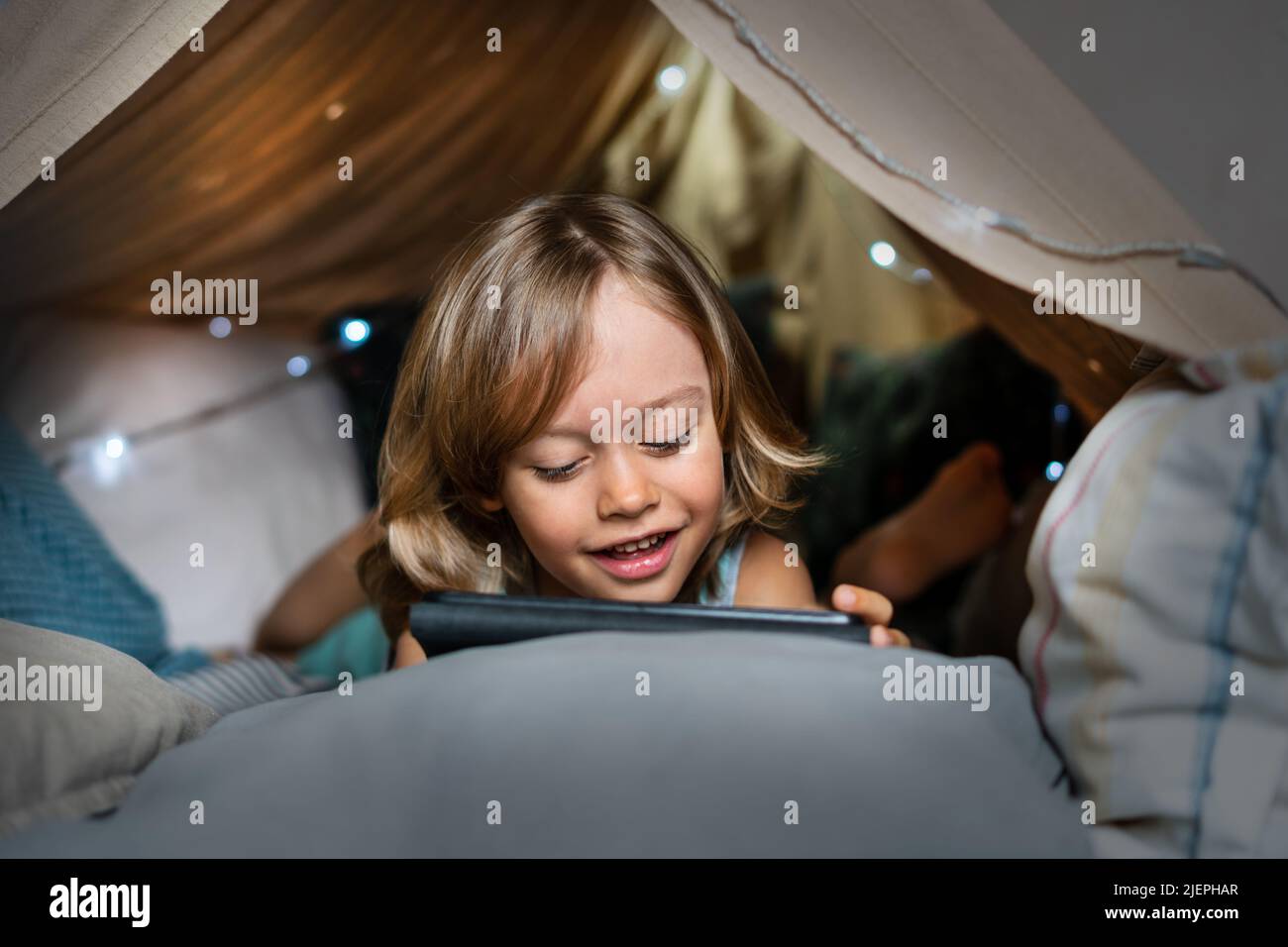 Happy cute little 6 year old boy having fun playing in teepee tent. Child using digital tablet watching cartoons or playing computer games lying in kid tent at home.  Stock Photo