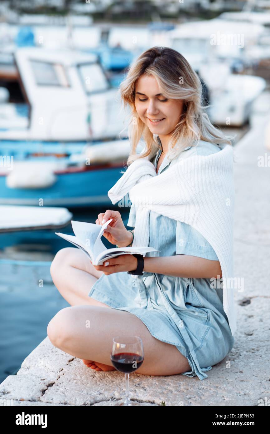 Happy young woman in light blue denim jumpsuit is sitting on pier with book in her hands, next to glass of red wine. Stock Photo