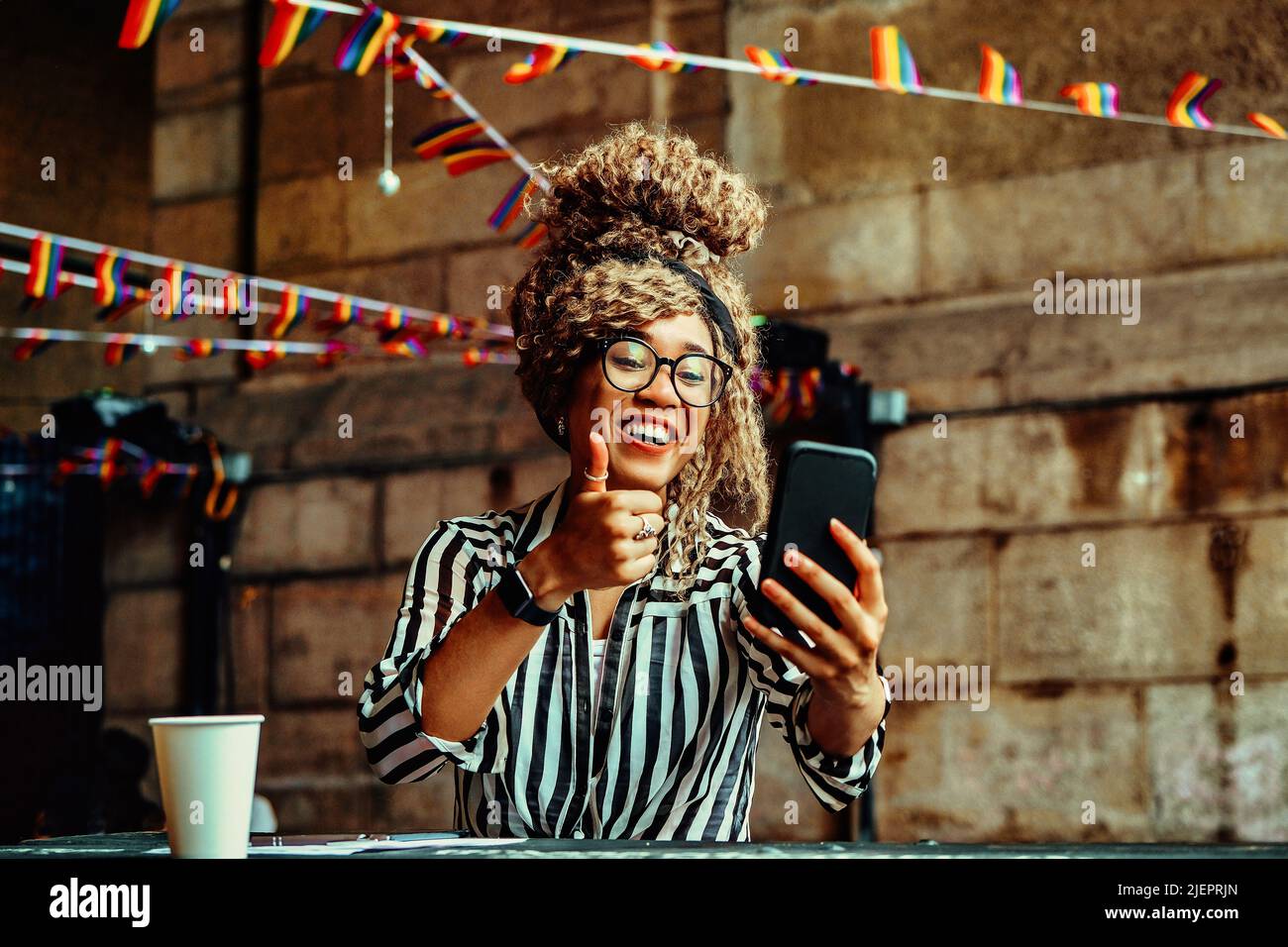 Portrait of smiling woman with afro hairstyle and eyeglasses on a phone video call with thumbs up sitting in a coffeehouse shot Stock Photo