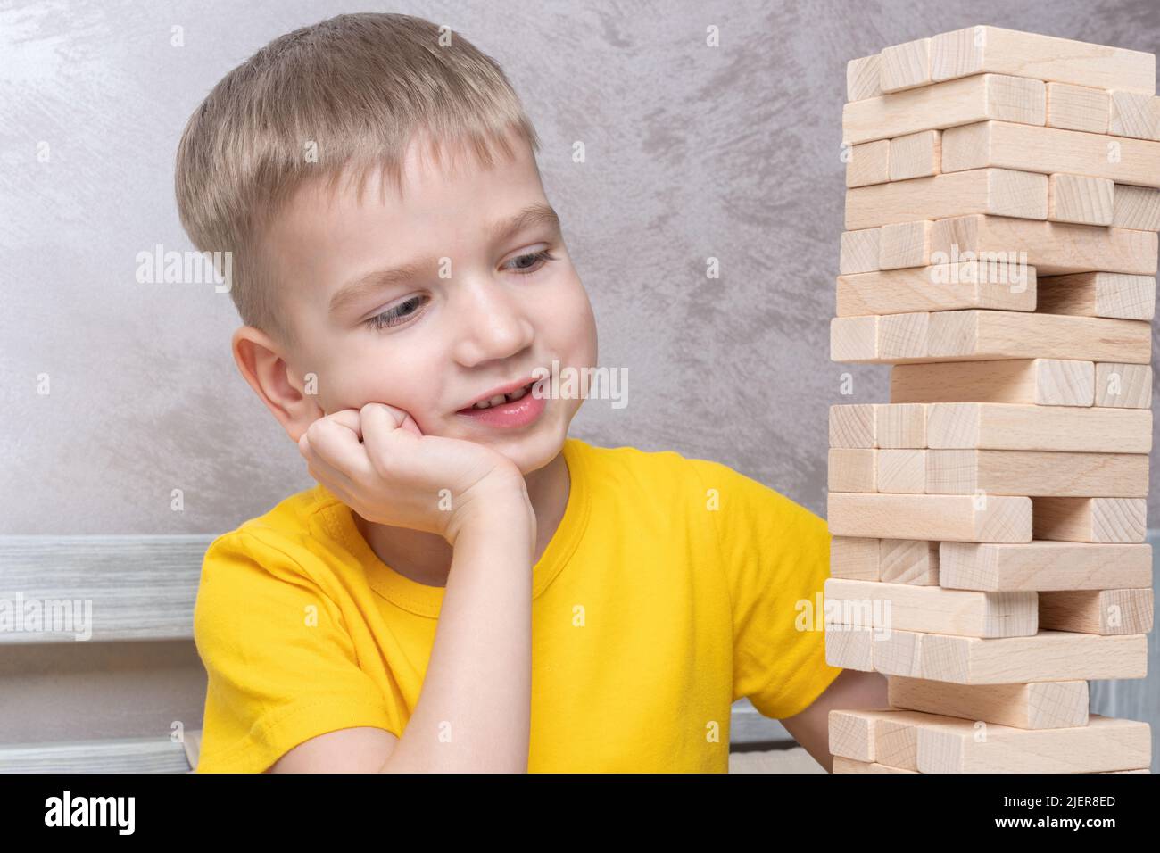 Interested happy little blond boy playing board game taking bricks from wooden tower keeping balance having fun together at home. Stock Photo