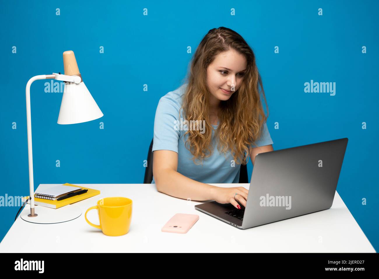 Young curly woman in a blue t-shirt studying from studio using laptop computer over a blue background. Serious beautiful young woman typing on laptop Stock Photo