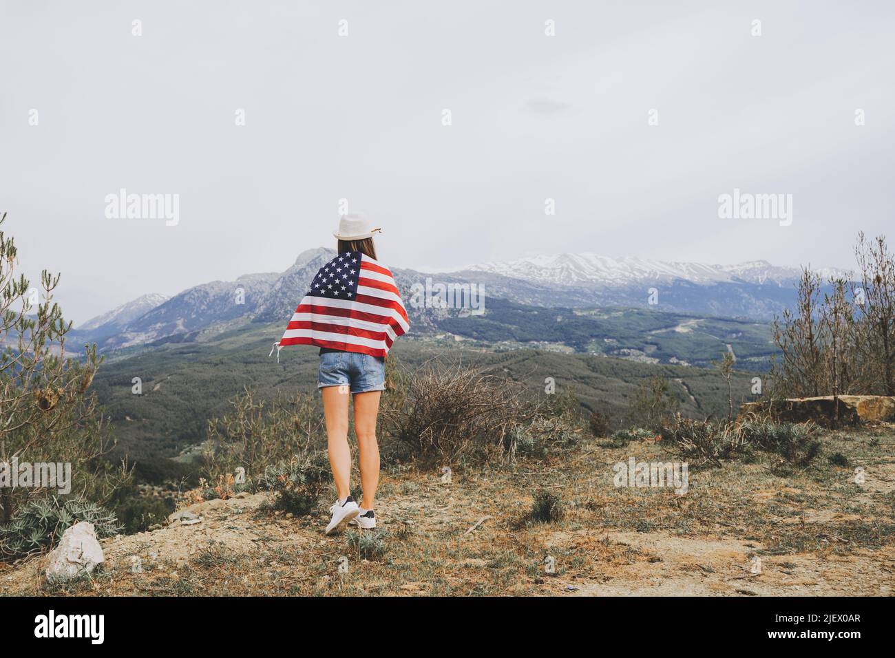 Young woman girl standing on rock cliff with US flag on her back and looking at mountains in background. Female traveller wearing American flag Stock Photo